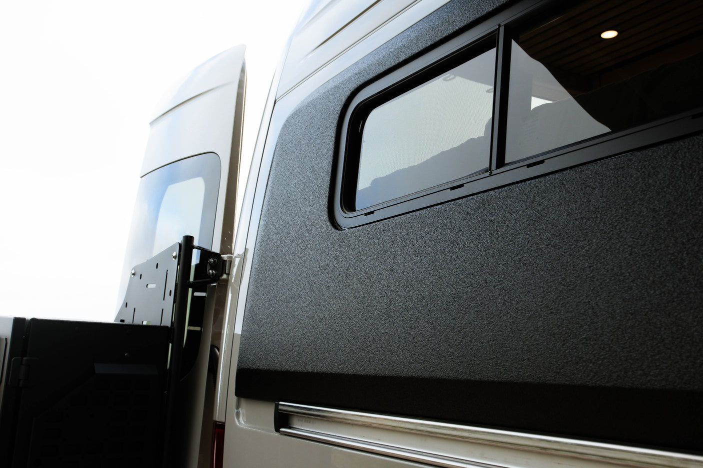 Side view of an RV rental: dark textured panel with an open window revealing the interior. Rear door and mounting bracket visible.