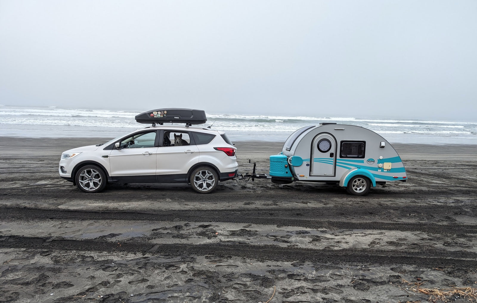 Enjoying an RV rental adventure! A white SUV with a dog inside tows a teardrop trailer on a misty beach by the ocean.|This light grey teardrop RV rental