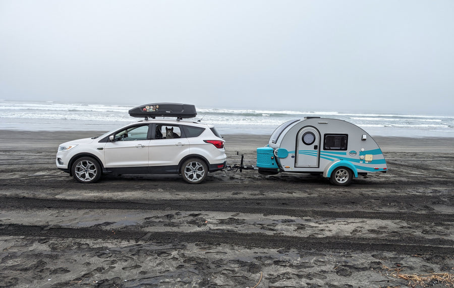 Enjoying an RV rental adventure! A white SUV with a dog inside tows a teardrop trailer on a misty beach by the ocean.|This light grey teardrop RV rental