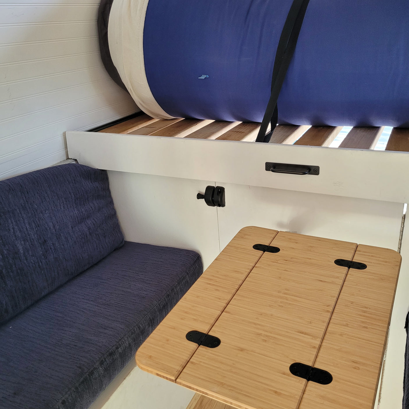Compact RV rental interior with blue cushioned seating, a fold-out bamboo table, and a raised sleeping area above.