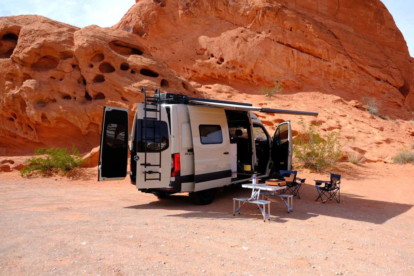 RV rental camper van set up for camping with table & chairs in a desert landscape with dramatic red rock formations.