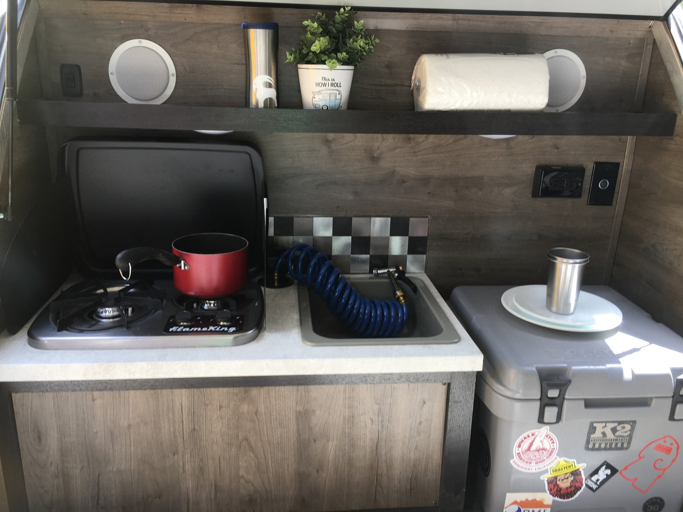 RV rental's compact kitchen setup featuring a gas stove, sink with pull-out hose, shelves, and a K2 cooler. — photo 5