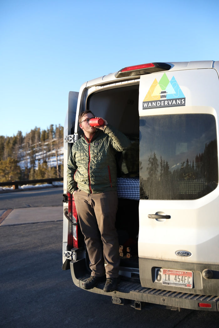 Man relaxing in a Wandervans RV rental. He lies in the Ford van's back with feet out, drinking from a red cup.