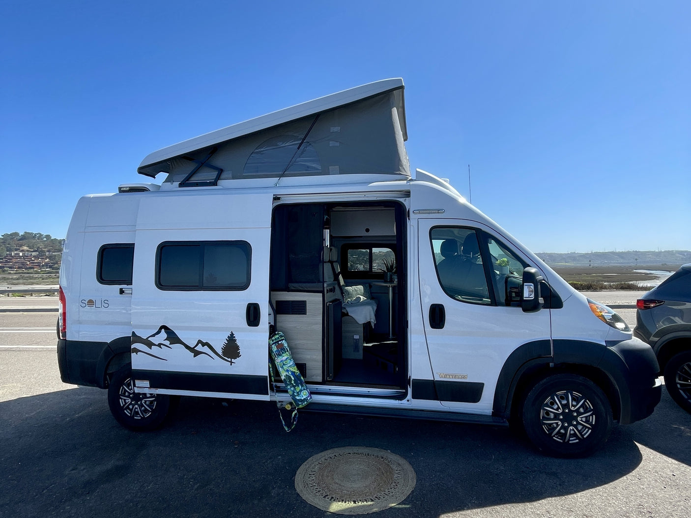 White Winnebago Solis camper van, great for rv rental, with pop-top extended & side door open. Skateboard & rug complete the mobile adventure scene.