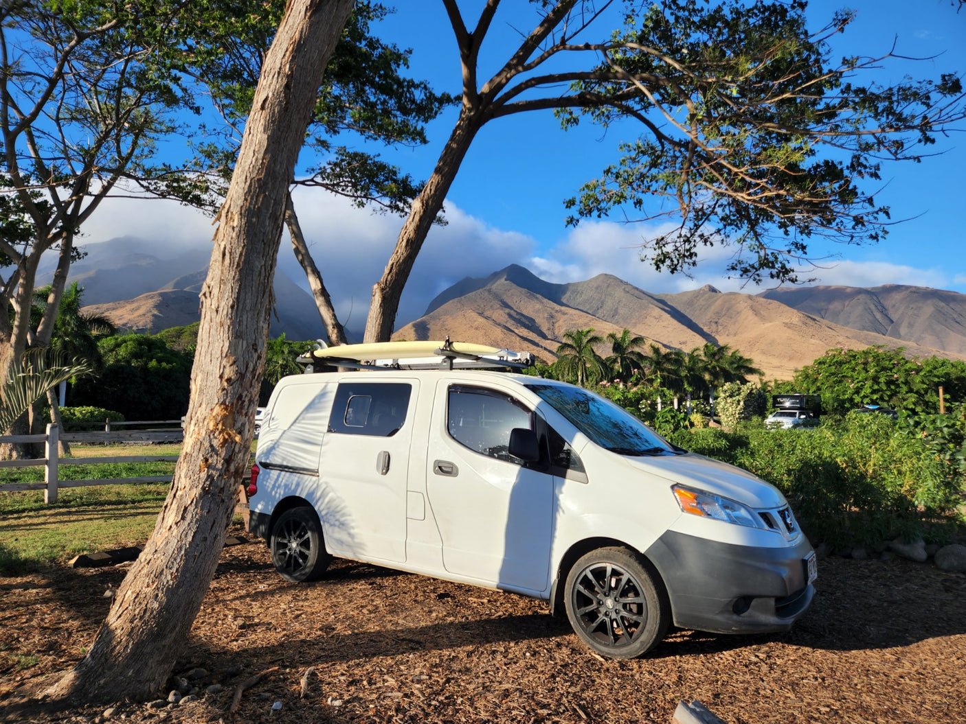 White van with surfboards on roof, parked under trees. Mountains and clouds create a scenic backdrop for this RV rental.