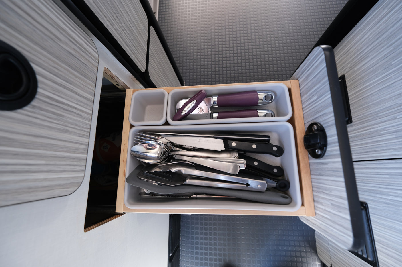 Compact RV rental kitchen drawer with neatly organized utensils: knives, forks, spoons, can opener, and spatulas.