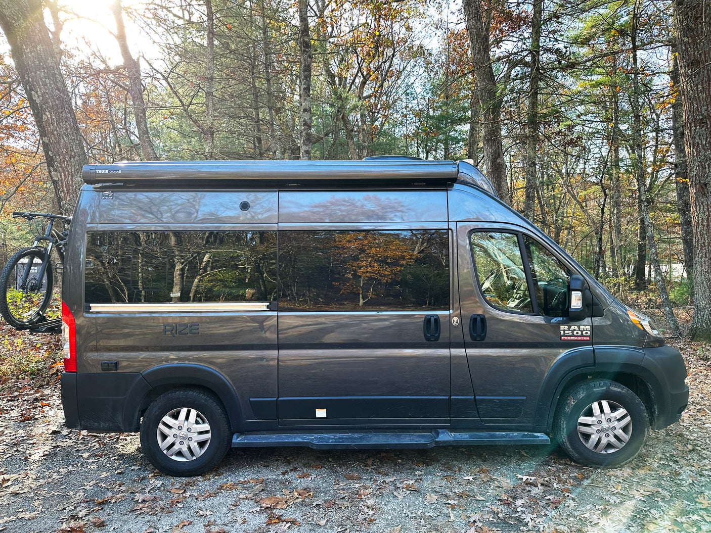 Explore nature with this grey Ram ProMaster camper van rv rental, featuring a bike rack, parked among sunlit trees.