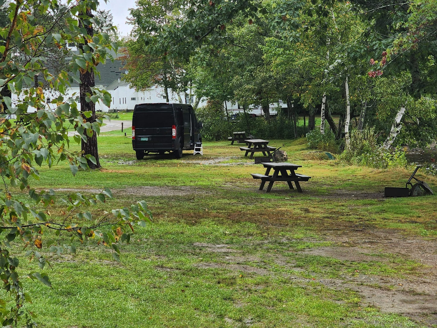 surrounded by trees and distant white buildings.|Black RV rental van parked lakeside on a grassy campsite with trees