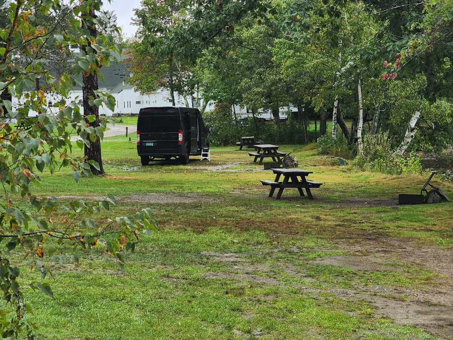 surrounded by trees and distant white buildings.|Black RV rental van parked lakeside on a grassy campsite with trees