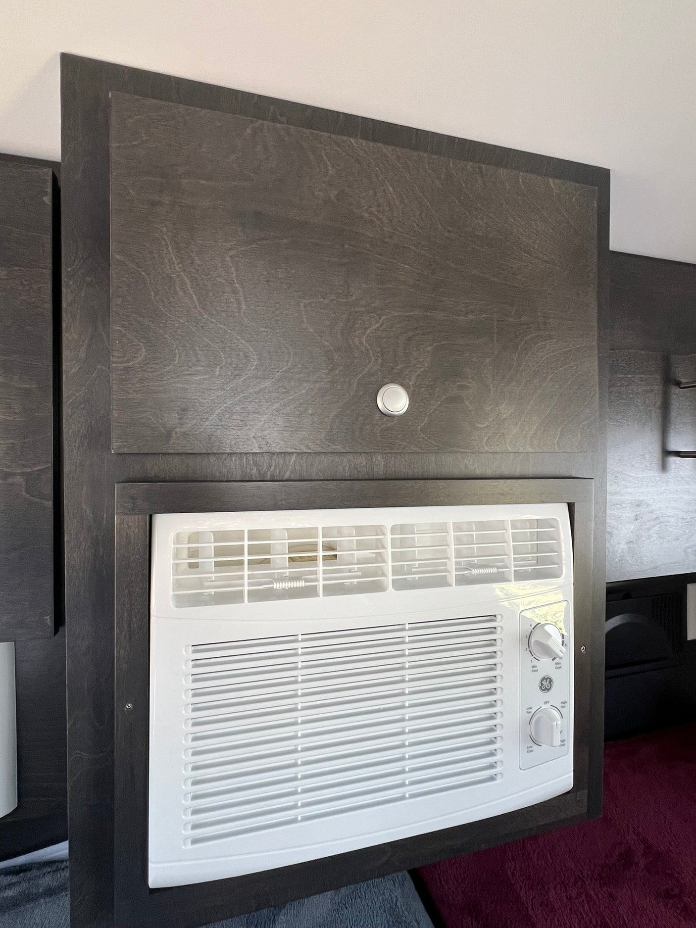RV rental comfort: A white GE air conditioner integrated into dark wood-grain cabinetry, with a storage cabinet above.