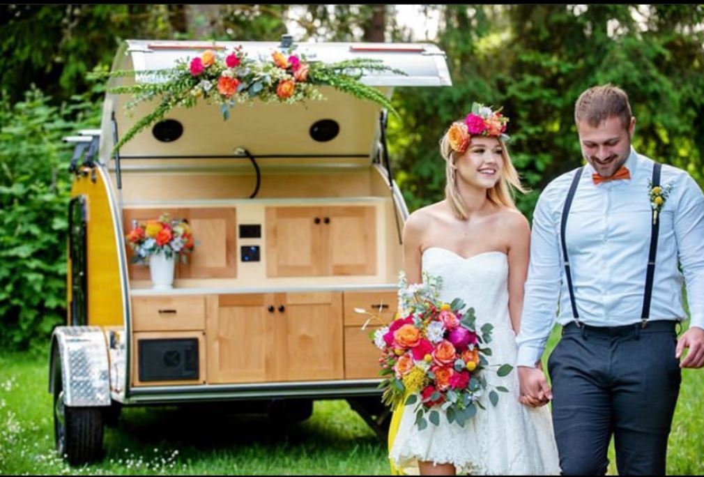 A happy couple holds hands by a yellow teardrop trailer with an open kitchen, decorated with flowers for their wedding. Consider this unique RV rental!