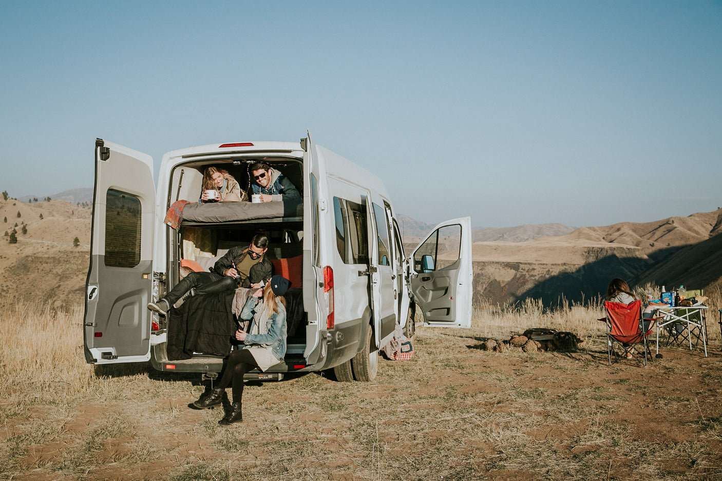 An RV rental offers ultimate freedom! Friends relax inside and outside a camper van, parked in a scenic mountain landscape.