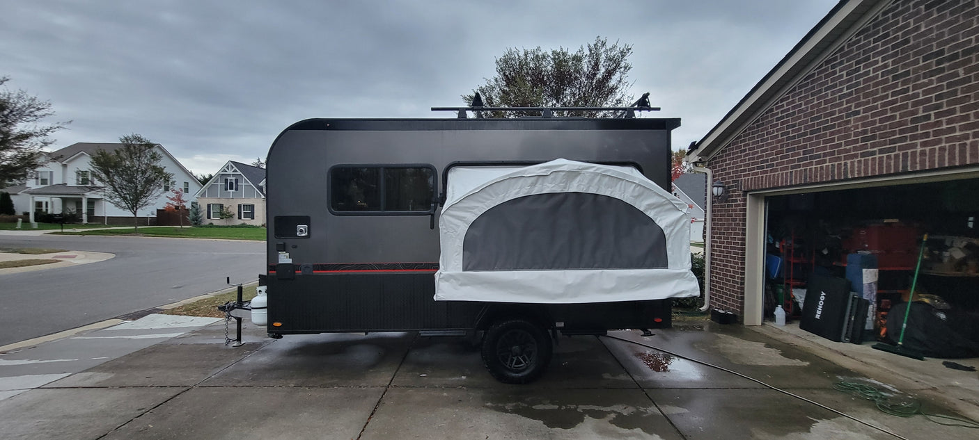 This dark gray travel trailer, an RV rental with a rear pop-out tent, is parked on a driveway, ready for adventure or a camping trip.