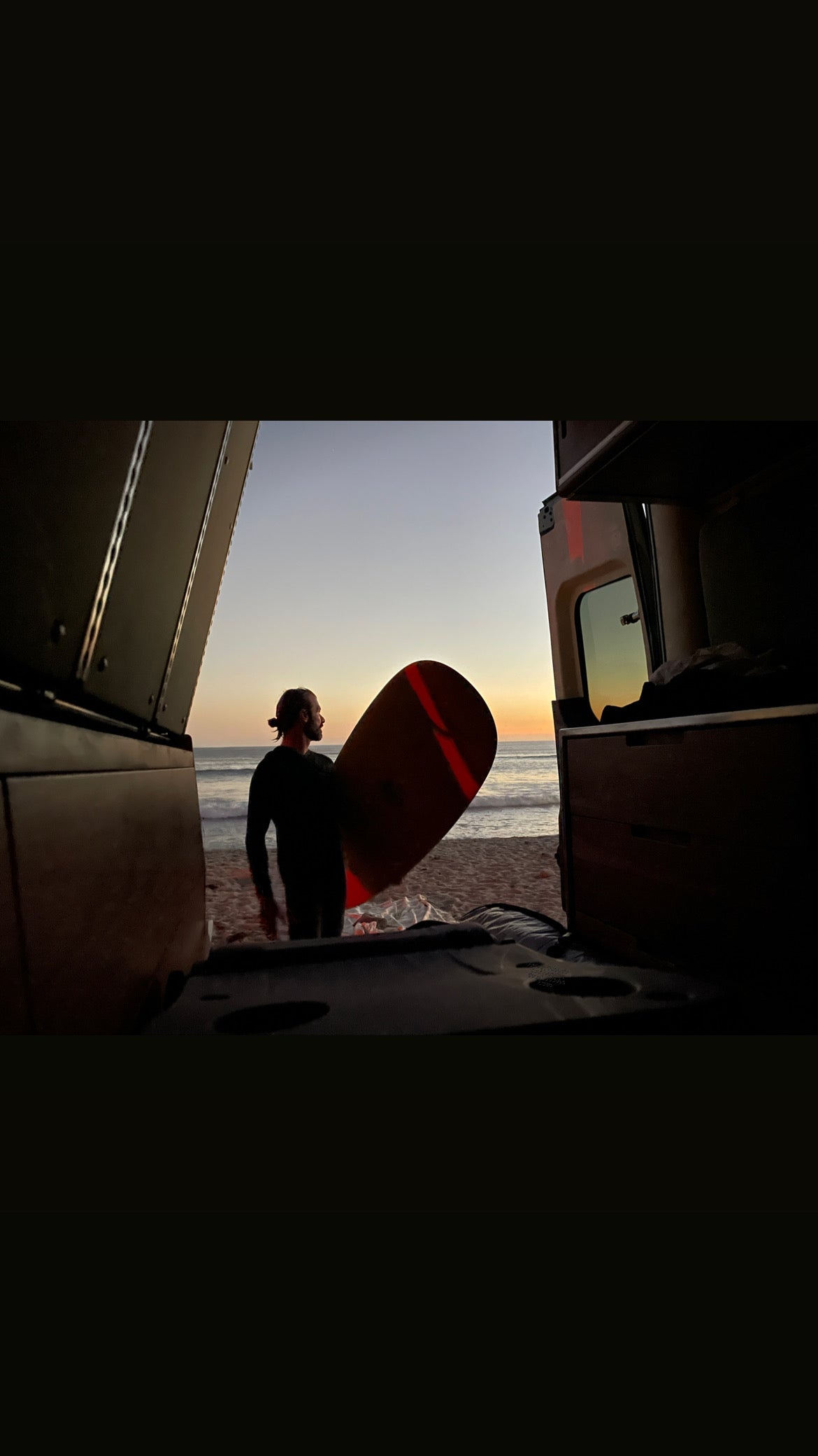 Surfer with board at sunset on beach, viewed from inside an RV rental. Ocean waves and colorful sky.