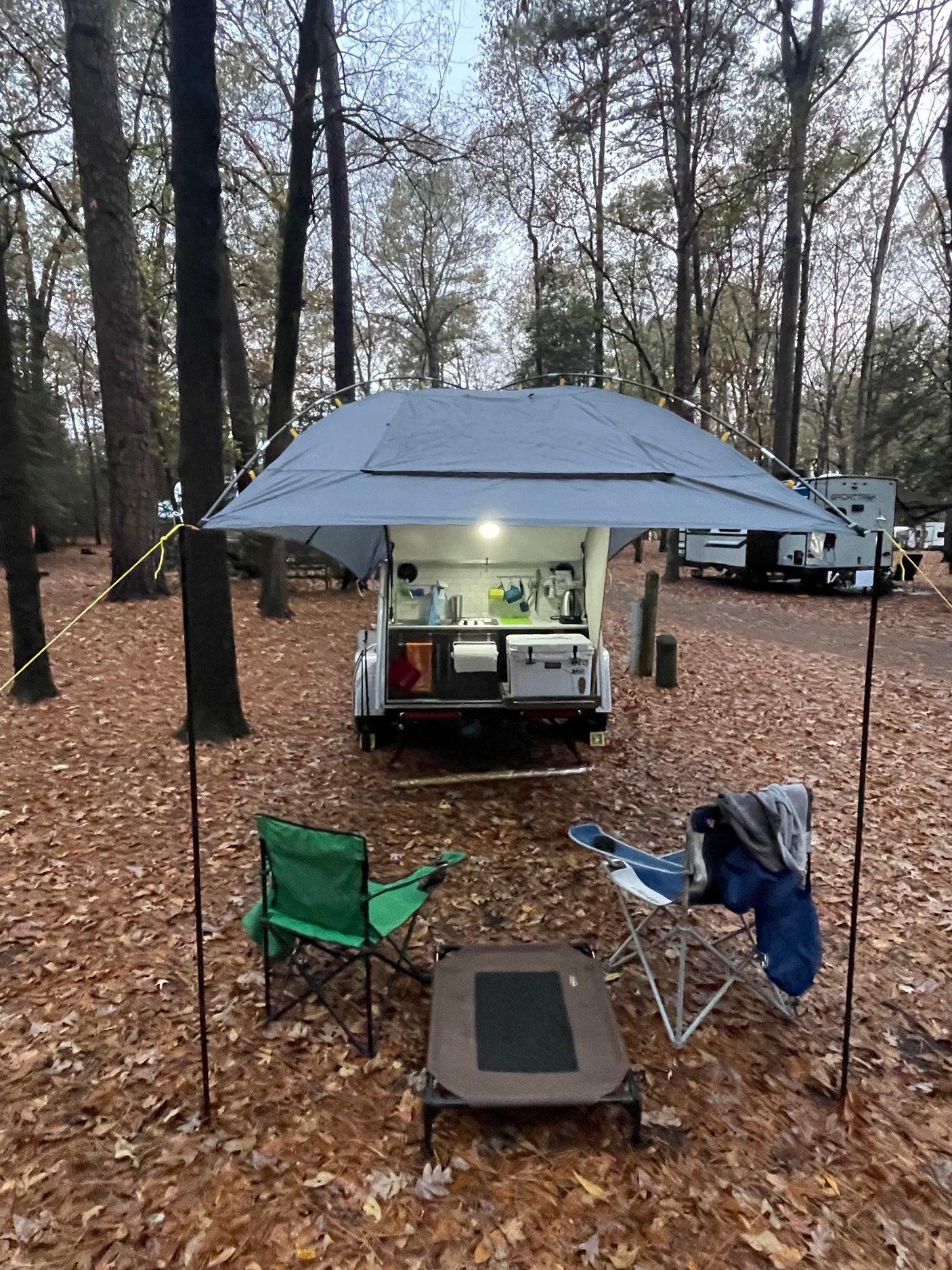 Cozy RV rental setup: a small camper with an outdoor kitchen & awning in a fall campsite, with chairs & pet bed.