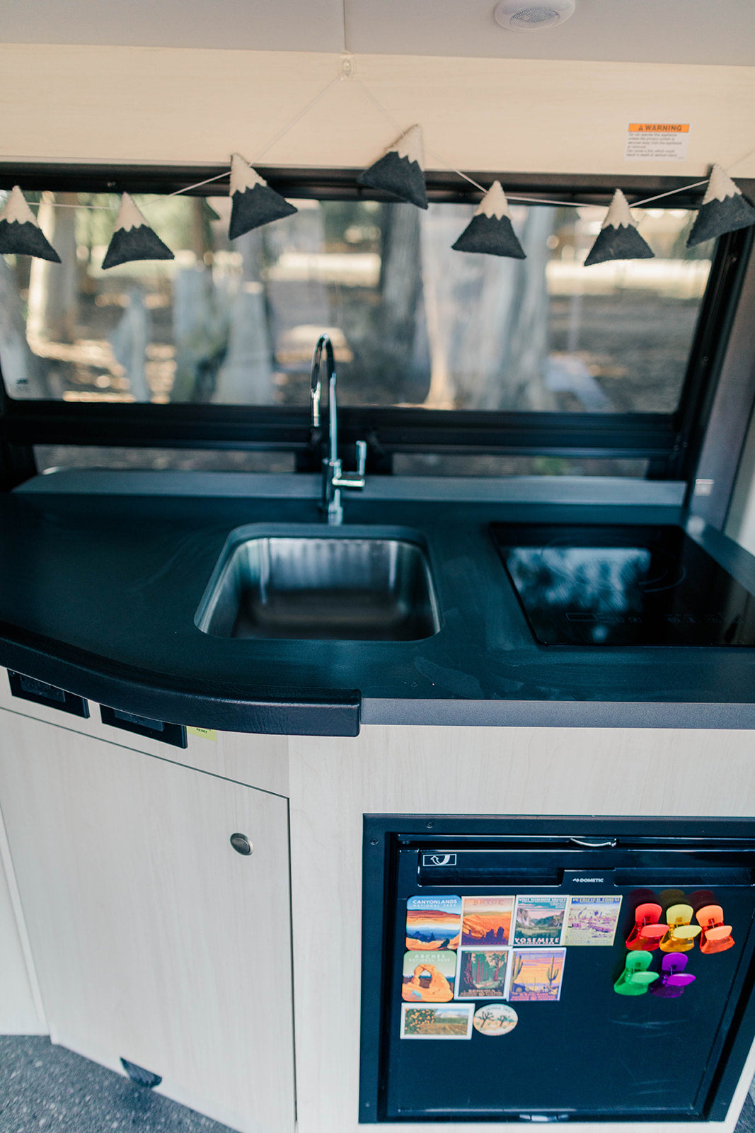 Inside an RV rental: a modern galley with a sink, induction cooktop, and mini-fridge adorned with national park magnets.
