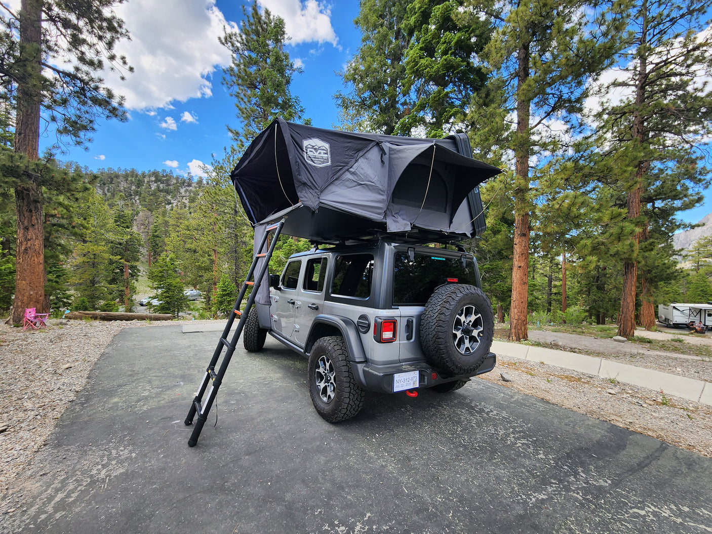 Experience nature with this gray Jeep Wrangler rv rental featuring a rooftop tent, set in a mountain campground among tall pines.