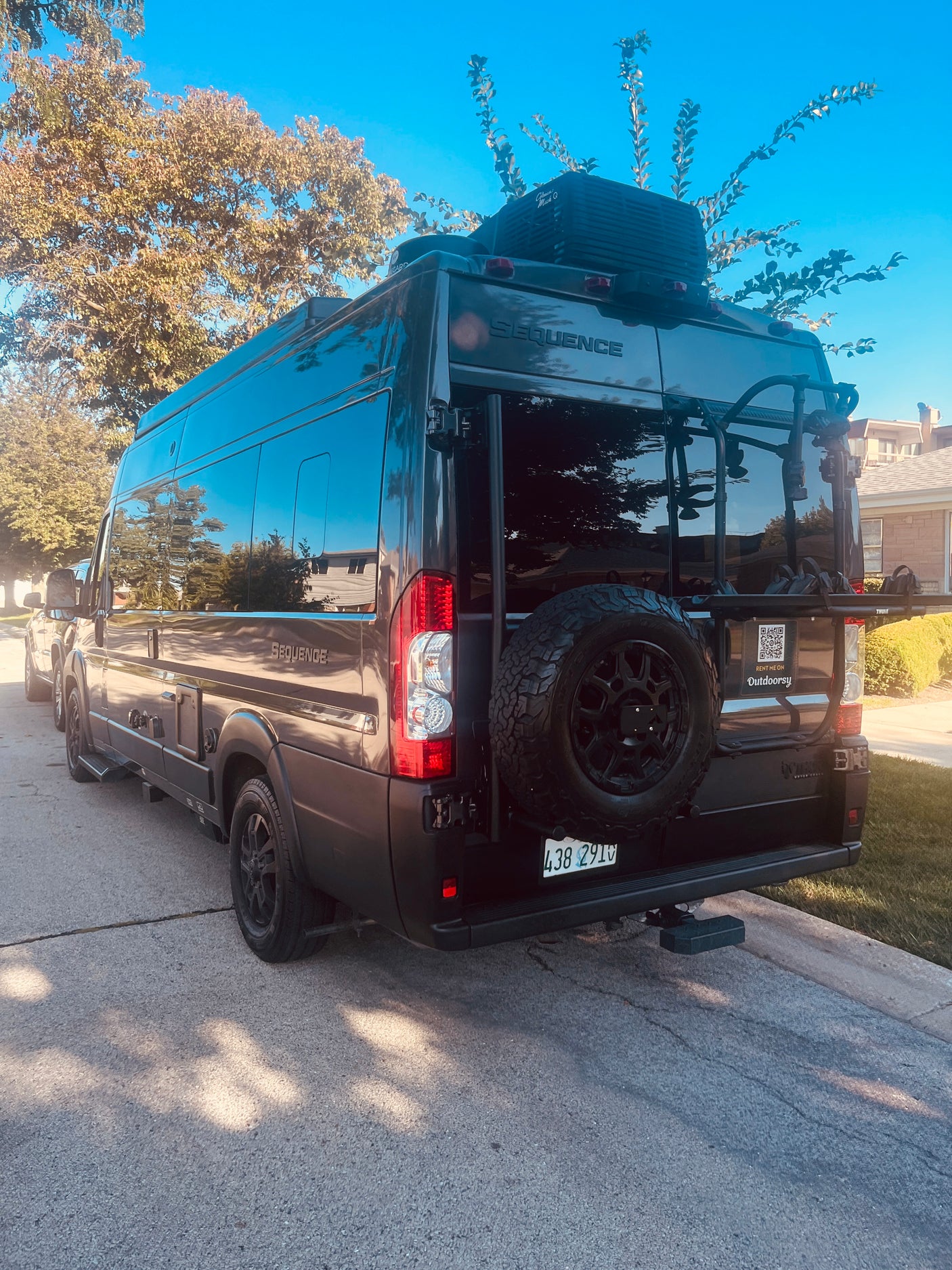 Dark gray Sequence camper van, ready as an RV rental, parked on asphalt with a spare tire and bike rack.