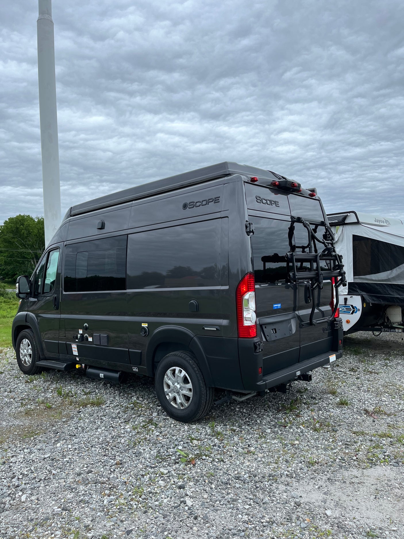 This dark gray SCOPE camper van, ideal for an RV rental adventure, features a bike rack and is parked on gravel near a pop-up camper.