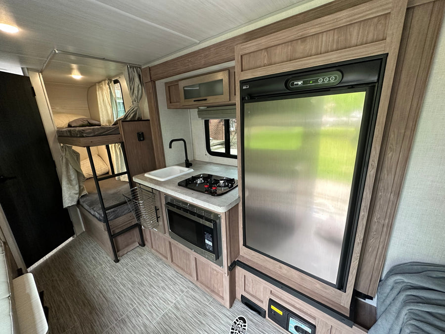 Interior of an RV rental showing a kitchen with sink, stove, microwave, and fridge, alongside comfy bunk beds.