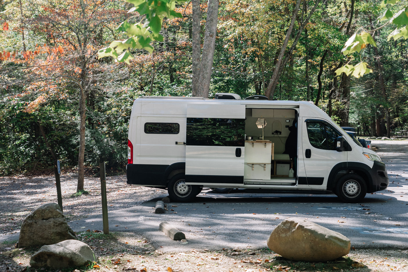White camper van (RV rental) parked in an autumn forest with its side door open, revealing a cozy interior for a nature getaway.