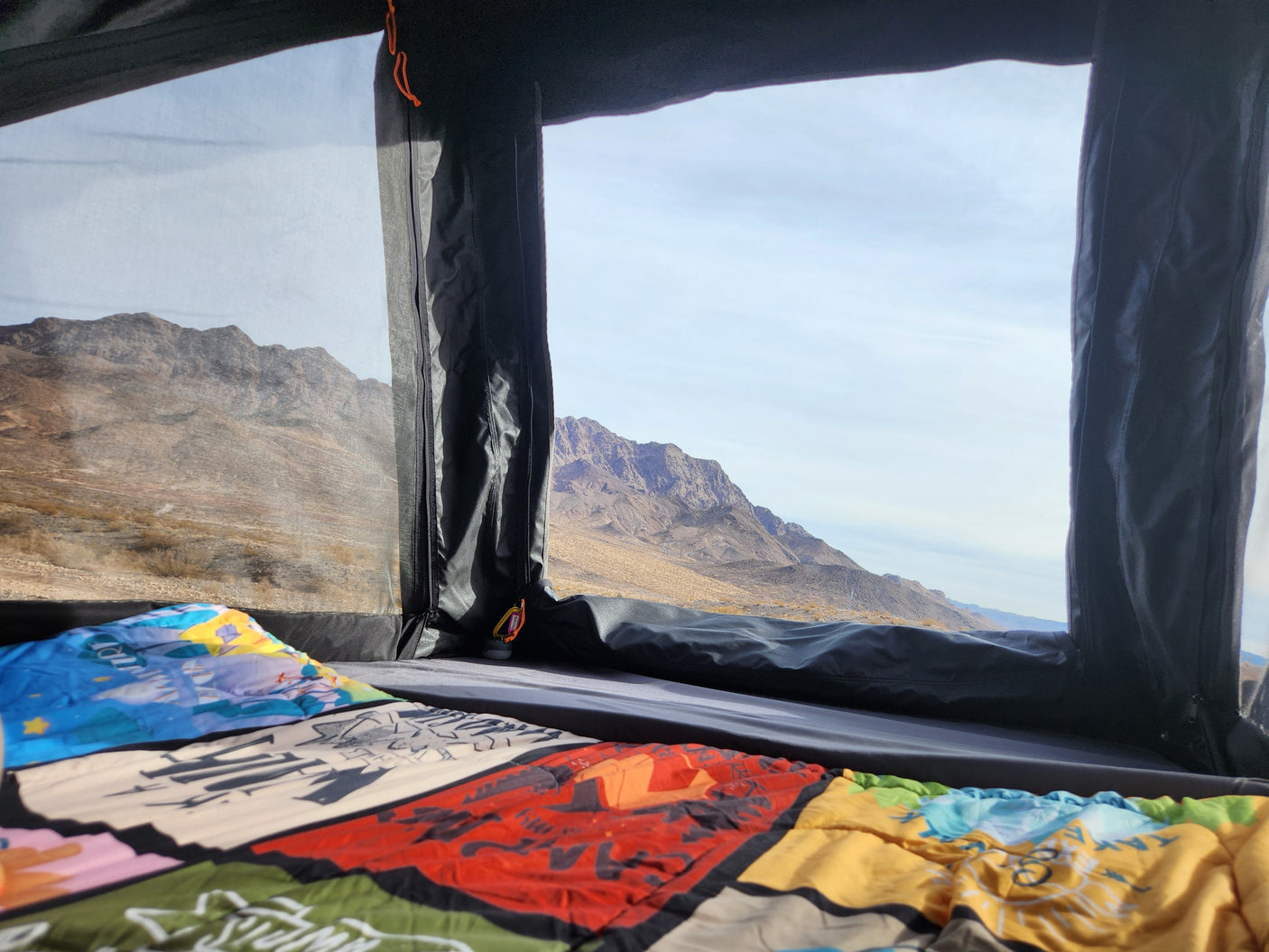 Inside an RV rental, a colorful blanket lies on the bed, offering a panoramic desert mountain view through the window.