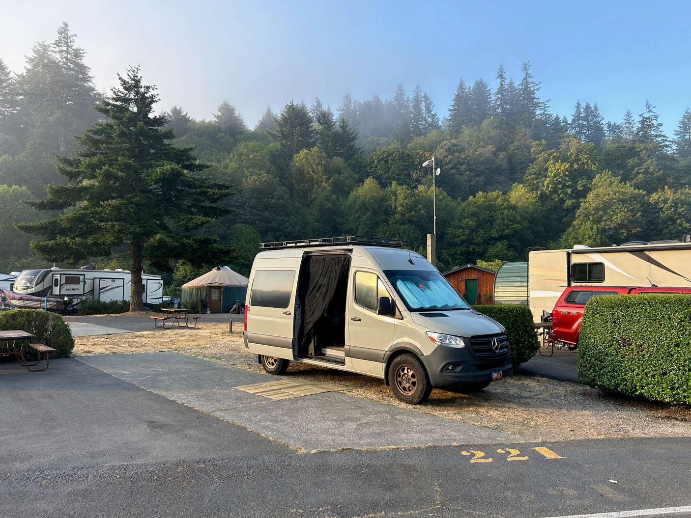 A light grey RV rental campervan parked at a scenic campground campsite 221, with misty evergreen trees and other RVs.