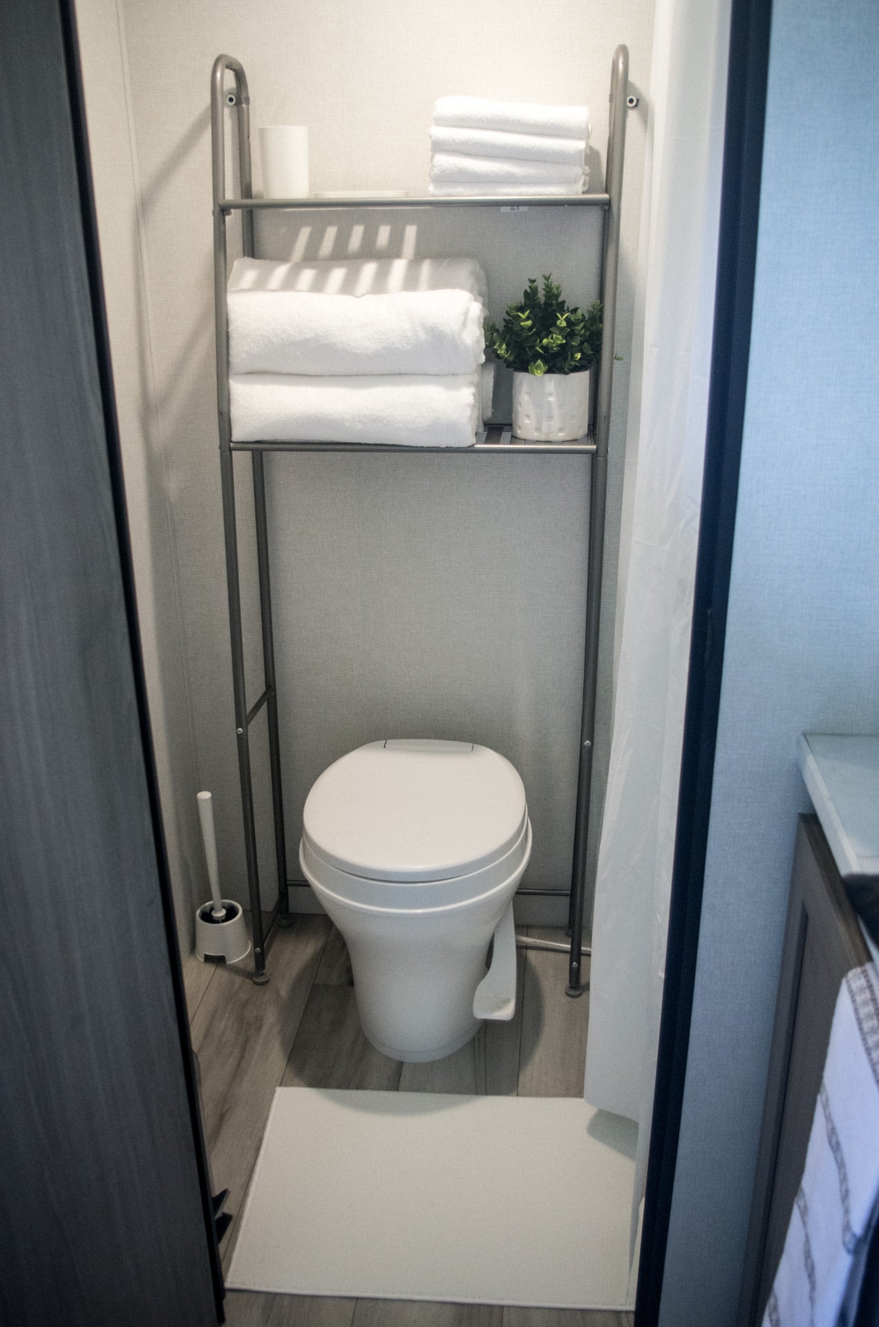 This RV rental bathroom features a compact toilet and over-toilet shelving with fresh towels and greenery. Essential travel amenities.