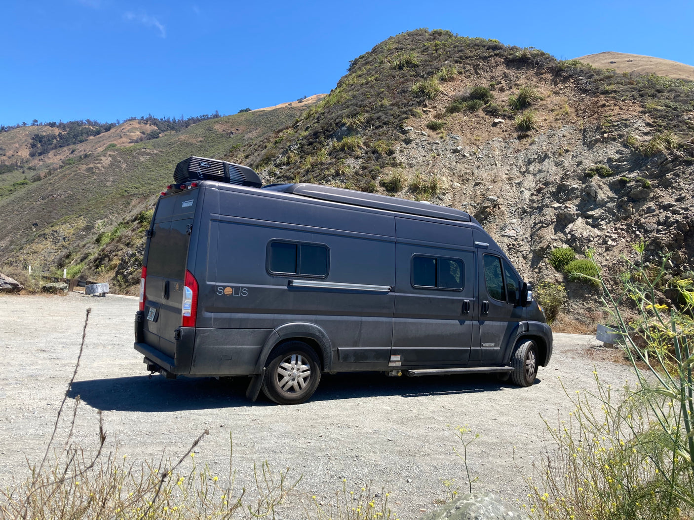 A dark grey Winnebago Solis camper van, available as an **rv rental**, parked on a gravel road with scenic mountains.