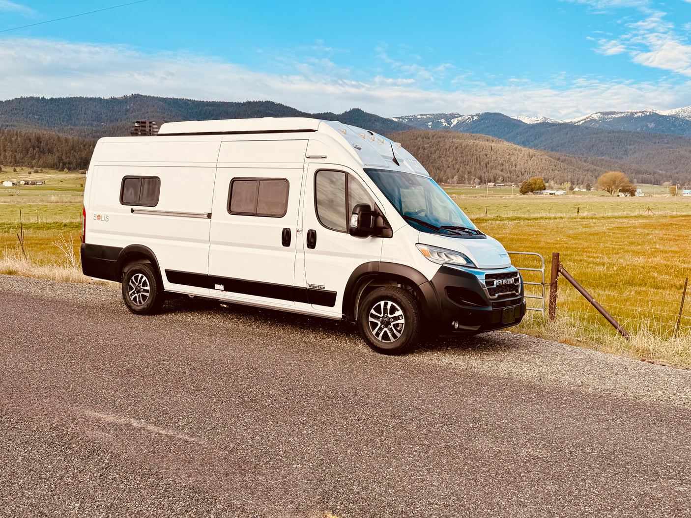 An RV rental - a white Winnebago Solis camper van - parked on a gravel road, with mountains & fields in the background.