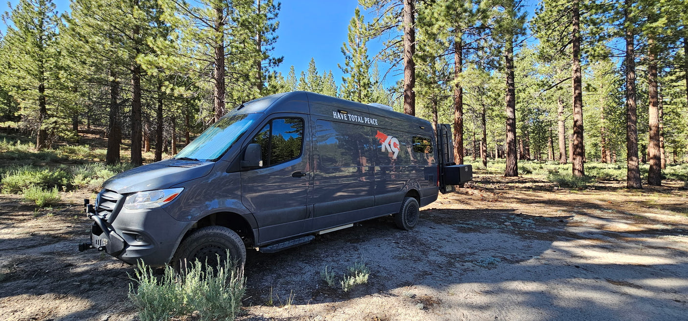 This dark grey Sprinter RV rental, with off-road features, is parked in a sunny pine forest, perfect for nature trips.