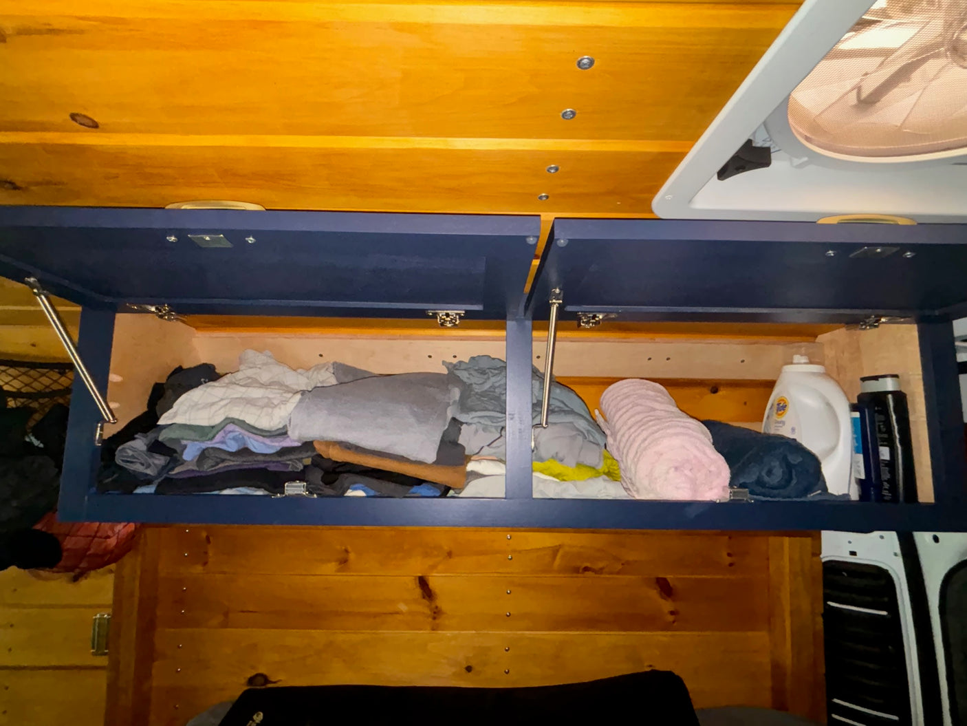 Open blue overhead cabinets in an RV rental reveal packed clothes, rolled towels, & essentials for travel.
