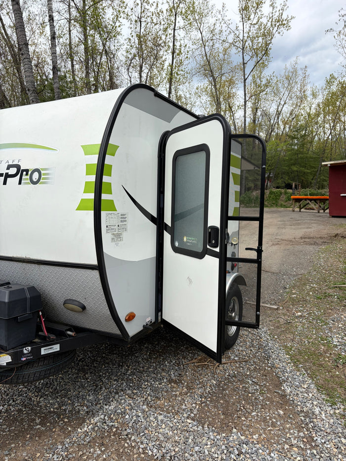 White and green travel trailer RV rental with door open, revealing screen. Parked on gravel, surrounded by trees for camping.