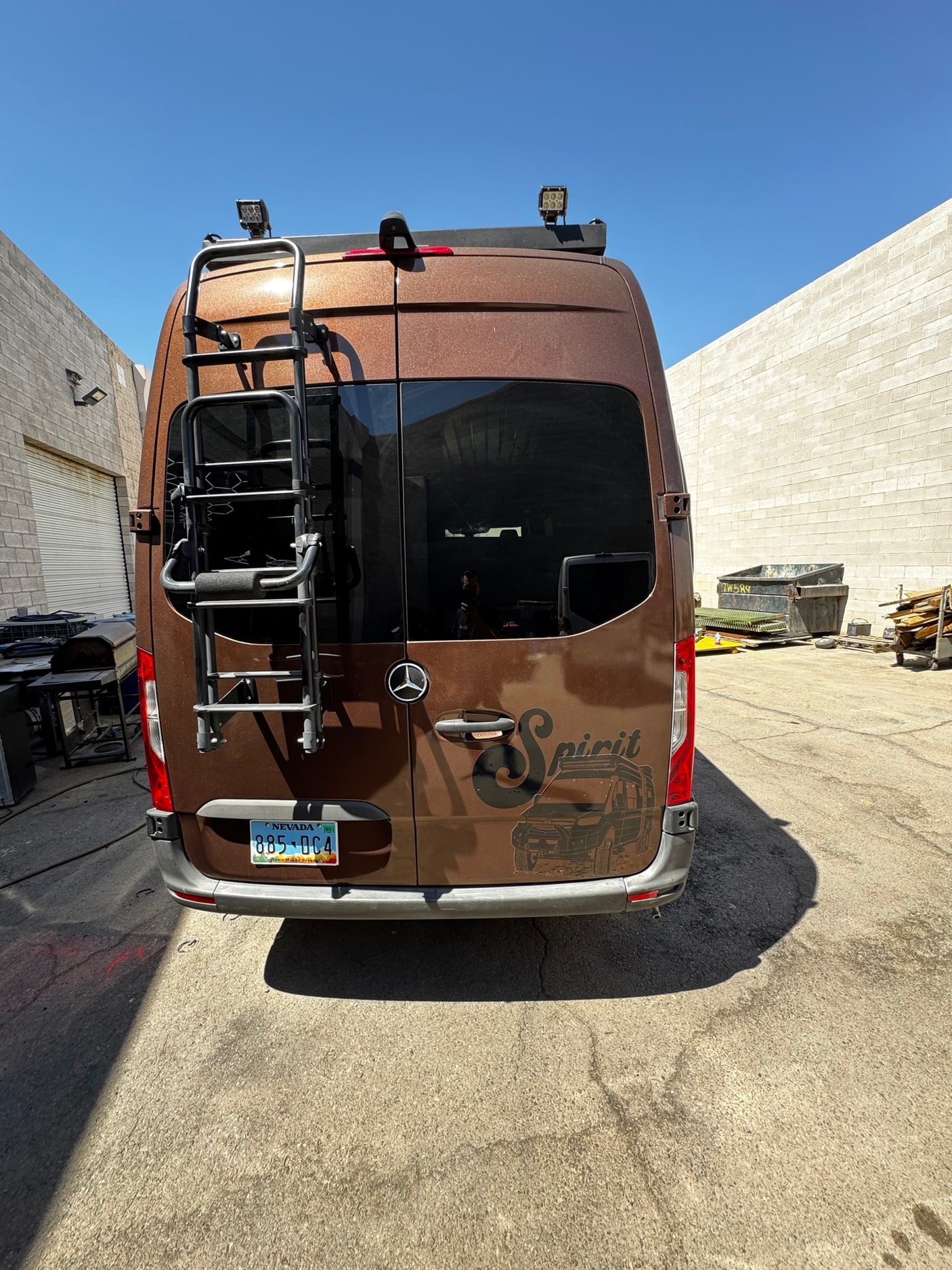 Rear of a brown Mercedes-Benz Sprinter RV rental van, customized with a ladder, roof lights, & 'Spirit' graphic. Parked outdoors.