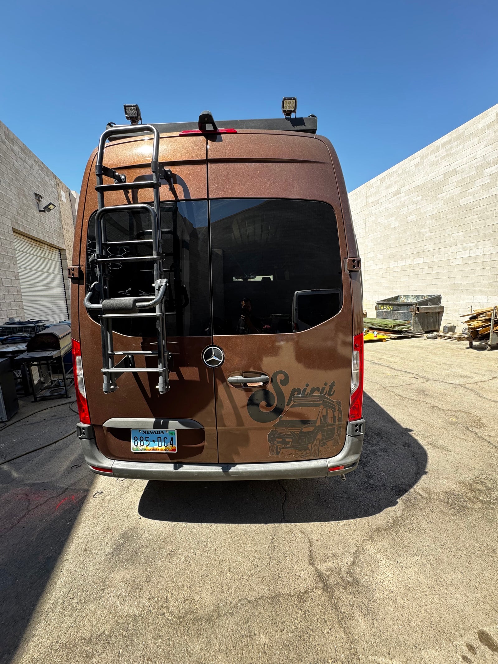 Rear of a brown Mercedes-Benz Sprinter RV rental van, customized with a ladder, roof lights, & 'Spirit' graphic. Parked outdoors.