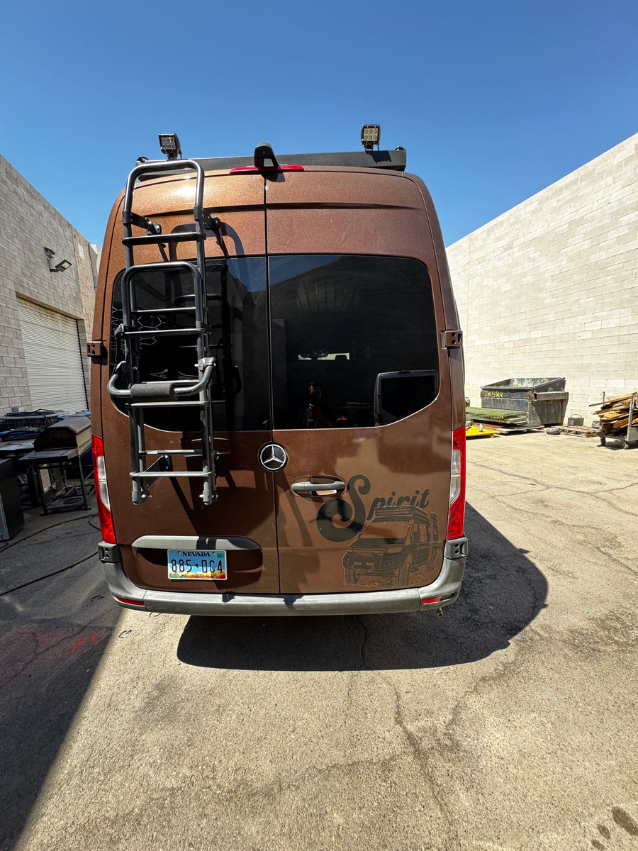 Rear of a brown Mercedes-Benz Sprinter RV rental van, customized with a ladder, roof lights, & 'Spirit' graphic. Parked outdoors.