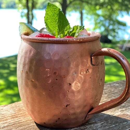 A cold drink with mint and lime in a hammered copper mug sits on a deck, overlooking a lake. Perfect after your RV rental adventure.