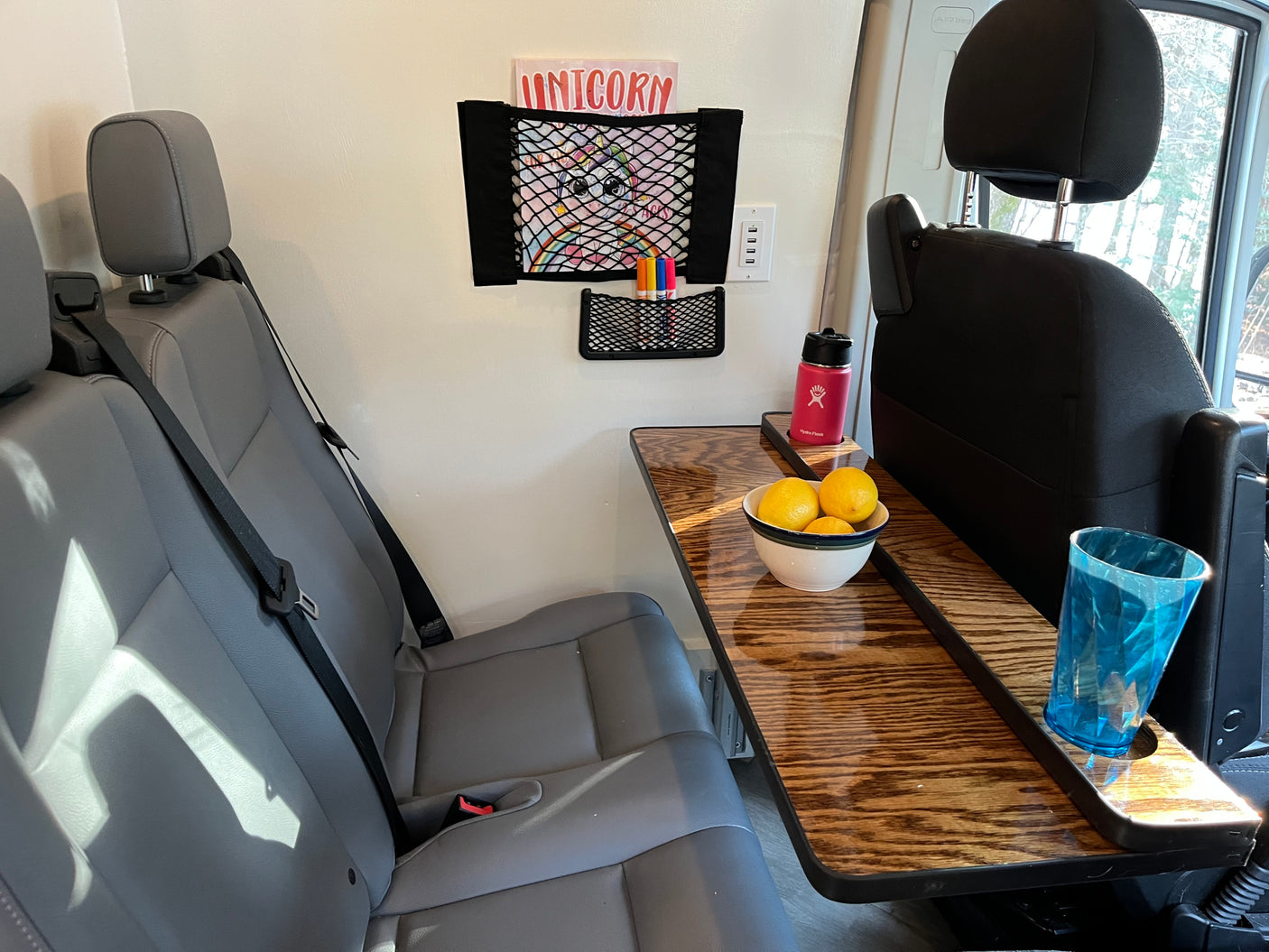 Cozy RV rental interior with passenger seats, a compact table holding lemons and drinks, plus wall storage for a book and chargers.