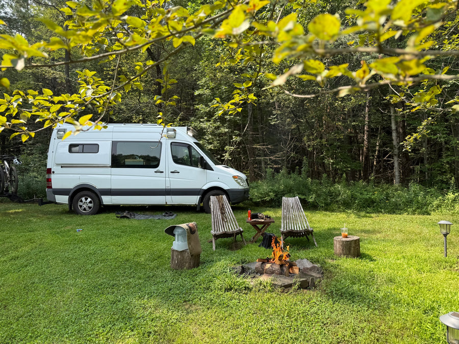 RV rental camper van with bikes, campfire, and chairs, nestled in a green forest setting.