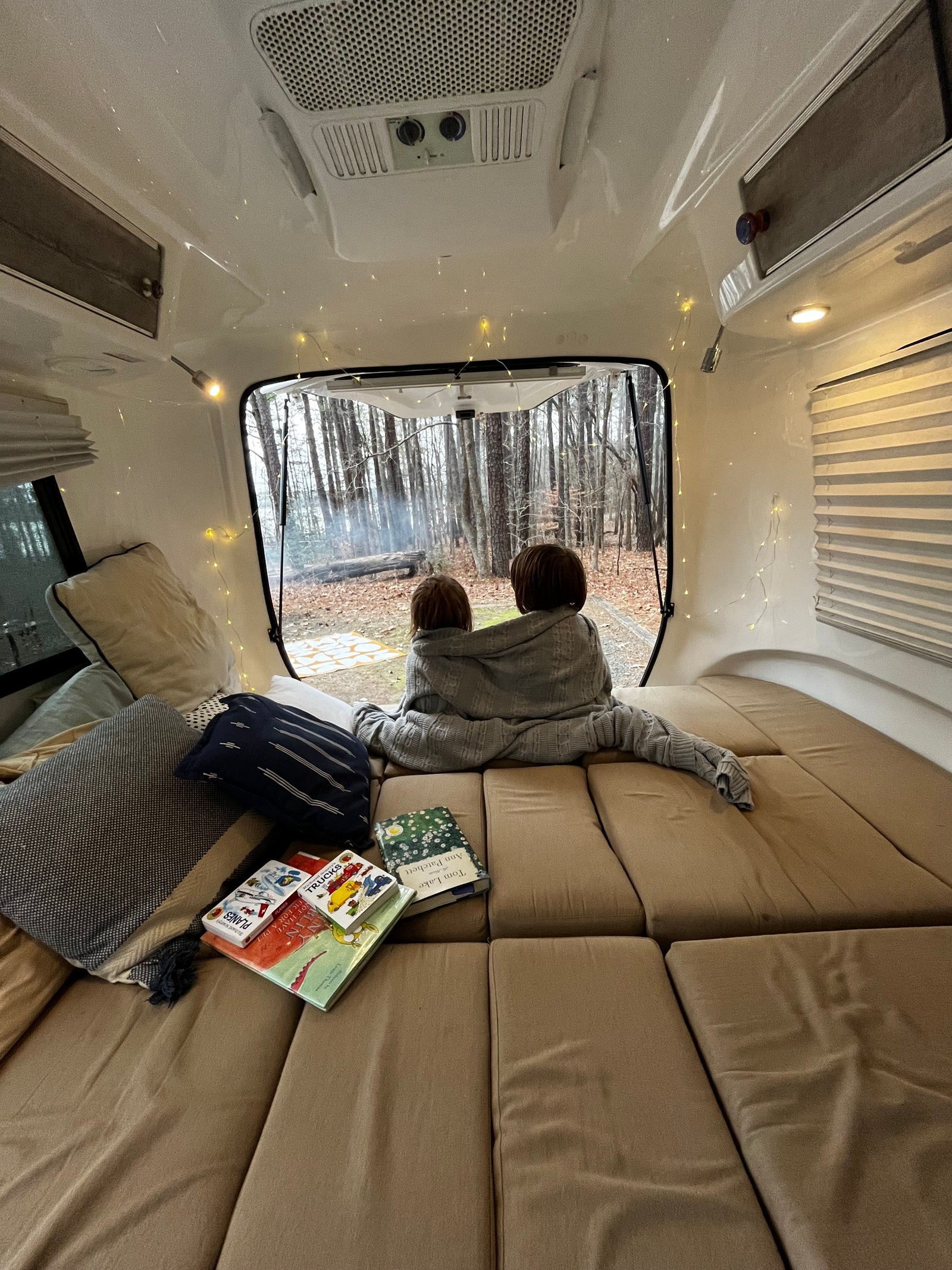 Inside a cozy **rv rental**: two children under a blanket watch a forest campfire. Books and string lights add to the warm atmosphere.