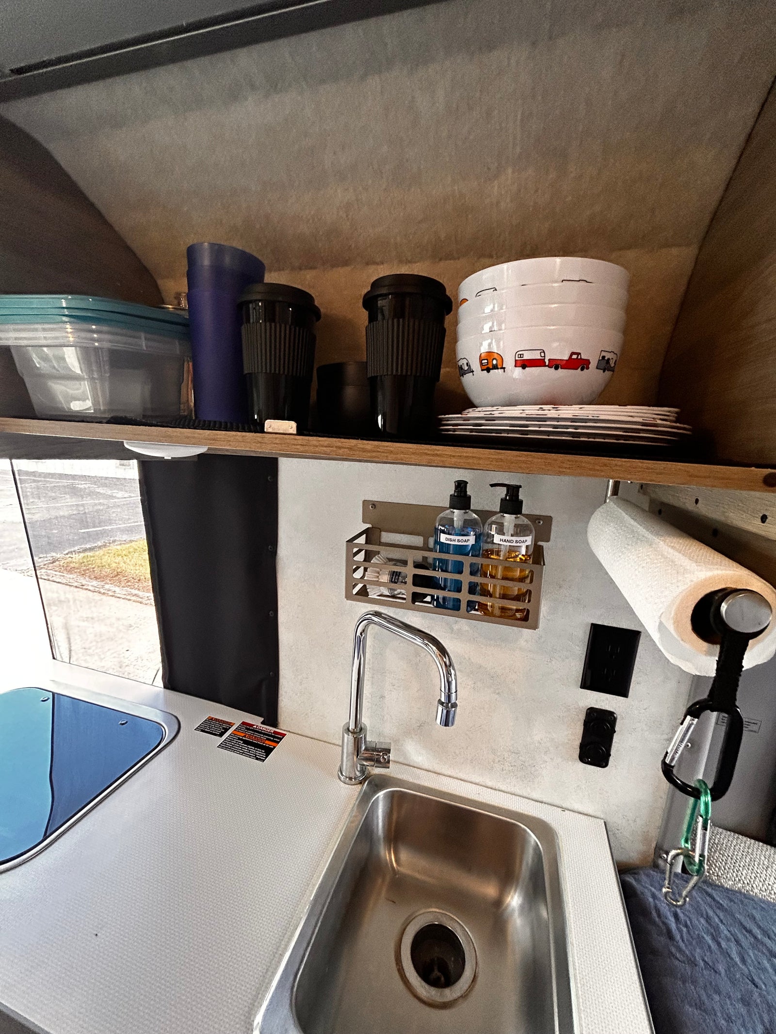 Inside an rv rental: a functional kitchenette with sink, covered stovetop, dishes, and cleaning supplies on shelves.
