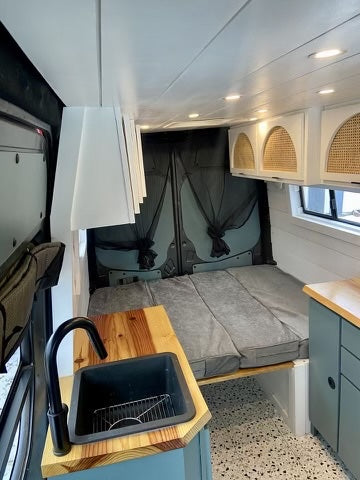 Stylish custom RV rental interior with a bed, sink, wooden counter, and cane-front cabinets for mobile living.