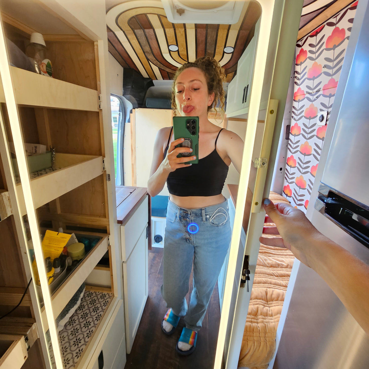 RV rental tour: Mirror selfie in custom van conversion. Woman shows unique wood ceiling, floral accents & compact storage.