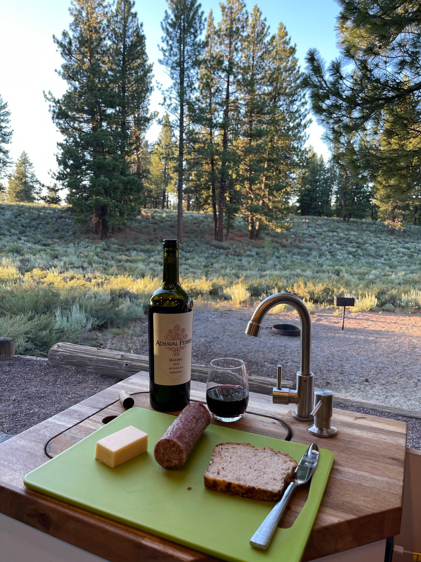 RV rental outdoor kitchen setup with wine, cheese, salami, bread, and forest views.