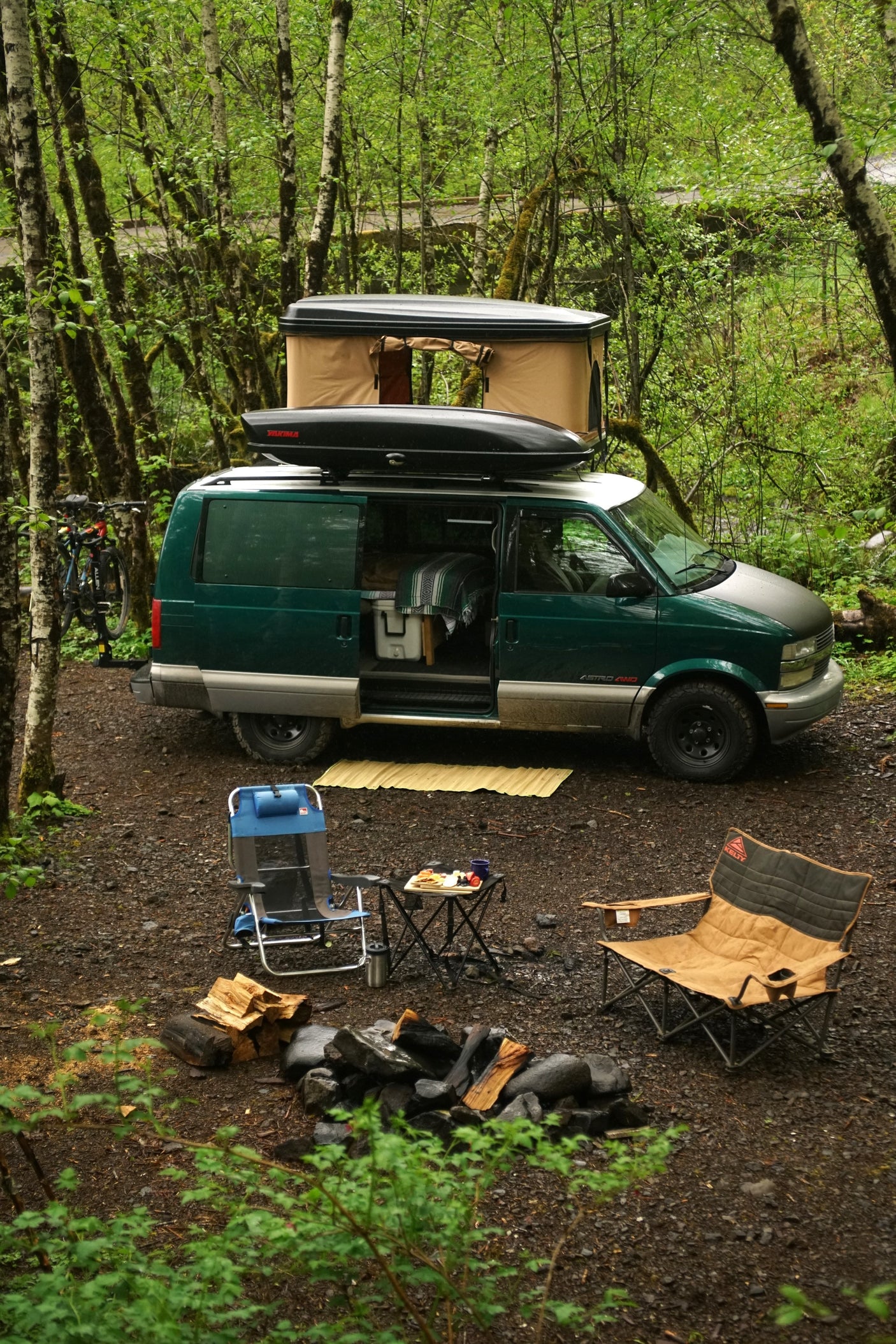 Van life adventure! This setup, like an RV rental, features a green Astro van with a rooftop tent, campfire, and chairs in a lush forest.