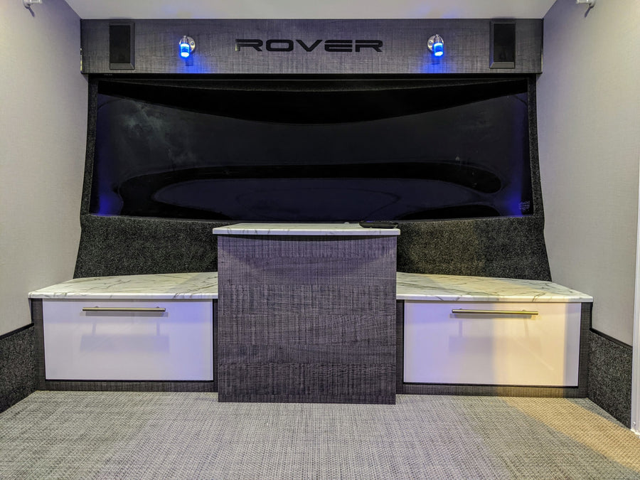 RV rental interior featuring a modern entertainment area with a large dark screen, "ROVER" branding, blue lights, and white/grey cabinets.