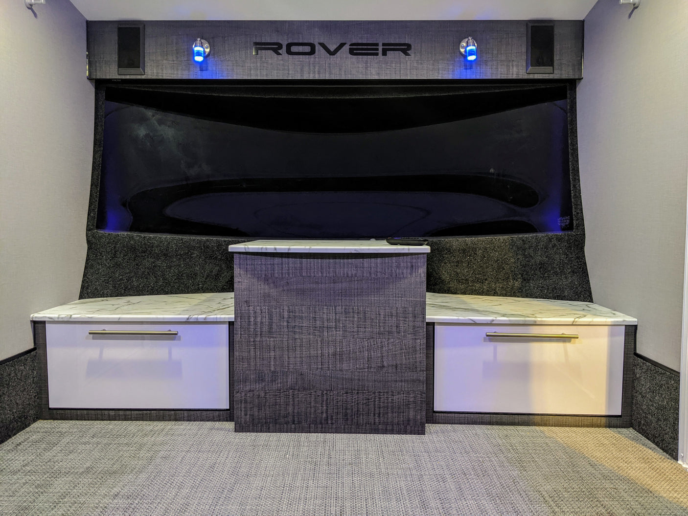 RV rental interior featuring a modern entertainment area with a large dark screen, "ROVER" branding, blue lights, and white/grey cabinets.