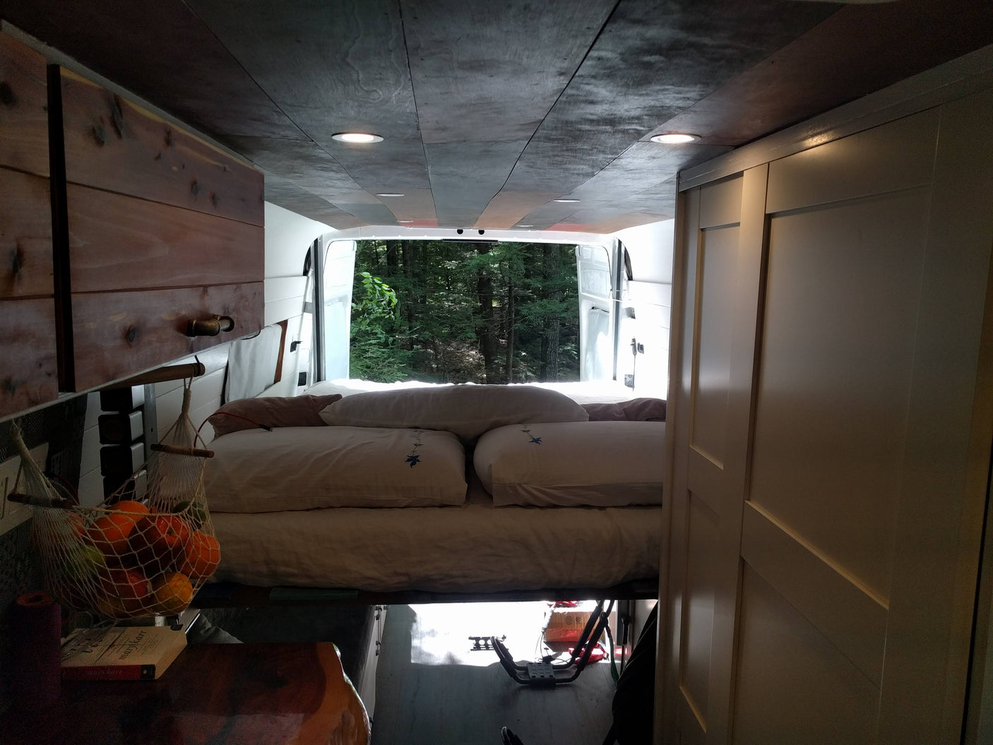 An RV rental's cozy interior features a bed, wood accents, and an expansive view of a verdant forest through open rear doors.