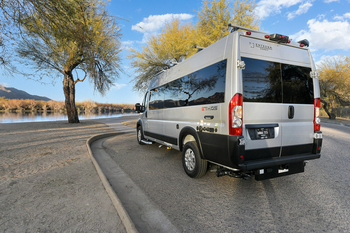 Silver Entegra Ethos RV rental parked by a scenic river under a blue sky, surrounded by trees. Perfect for adventure!