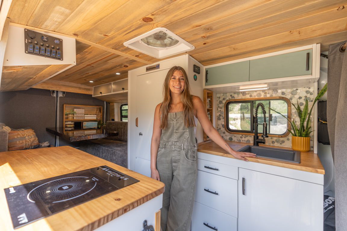 Smiling woman in her cozy RV rental, showcasing a modern camper van interior with kitchen, sleeping area, and wood ceiling.